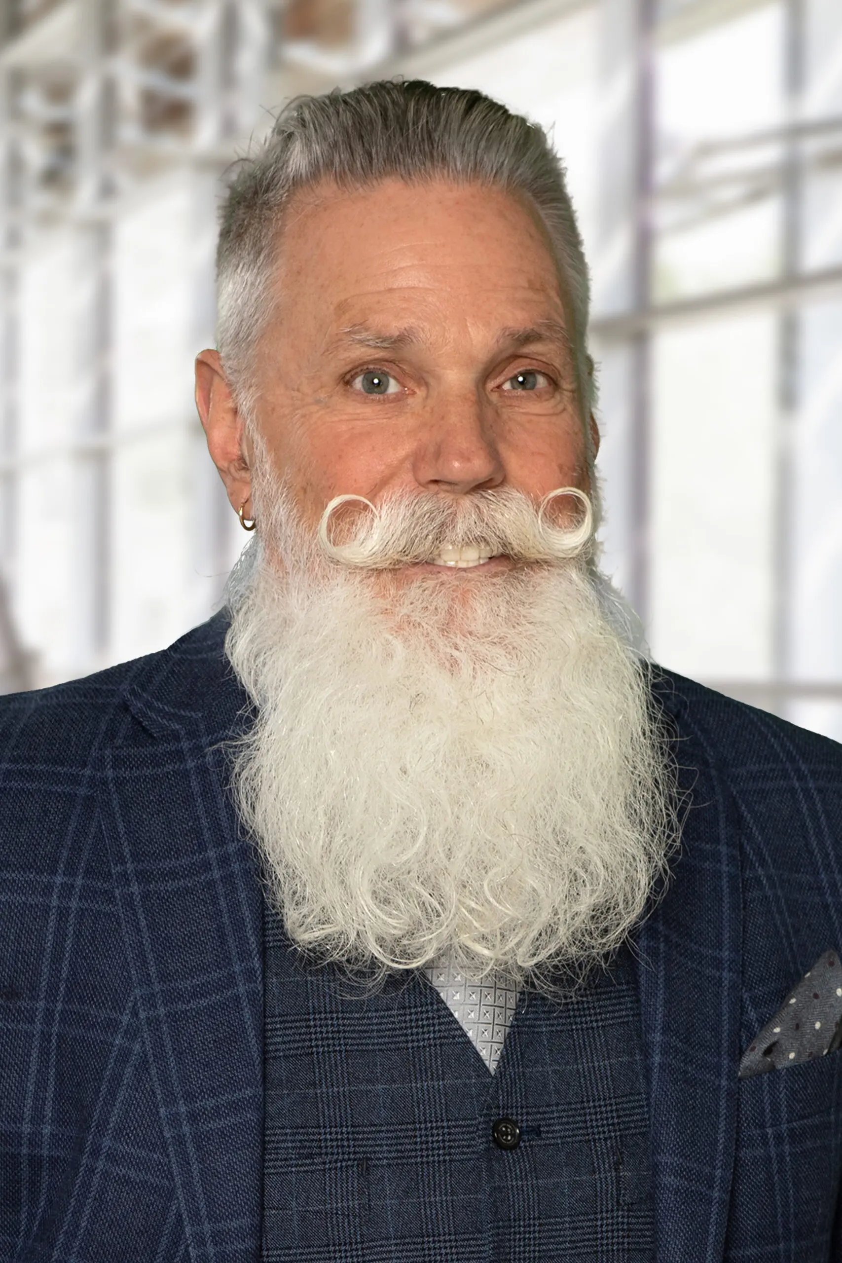 Elderly man with a long white beard and curled mustache in a checkered suit.