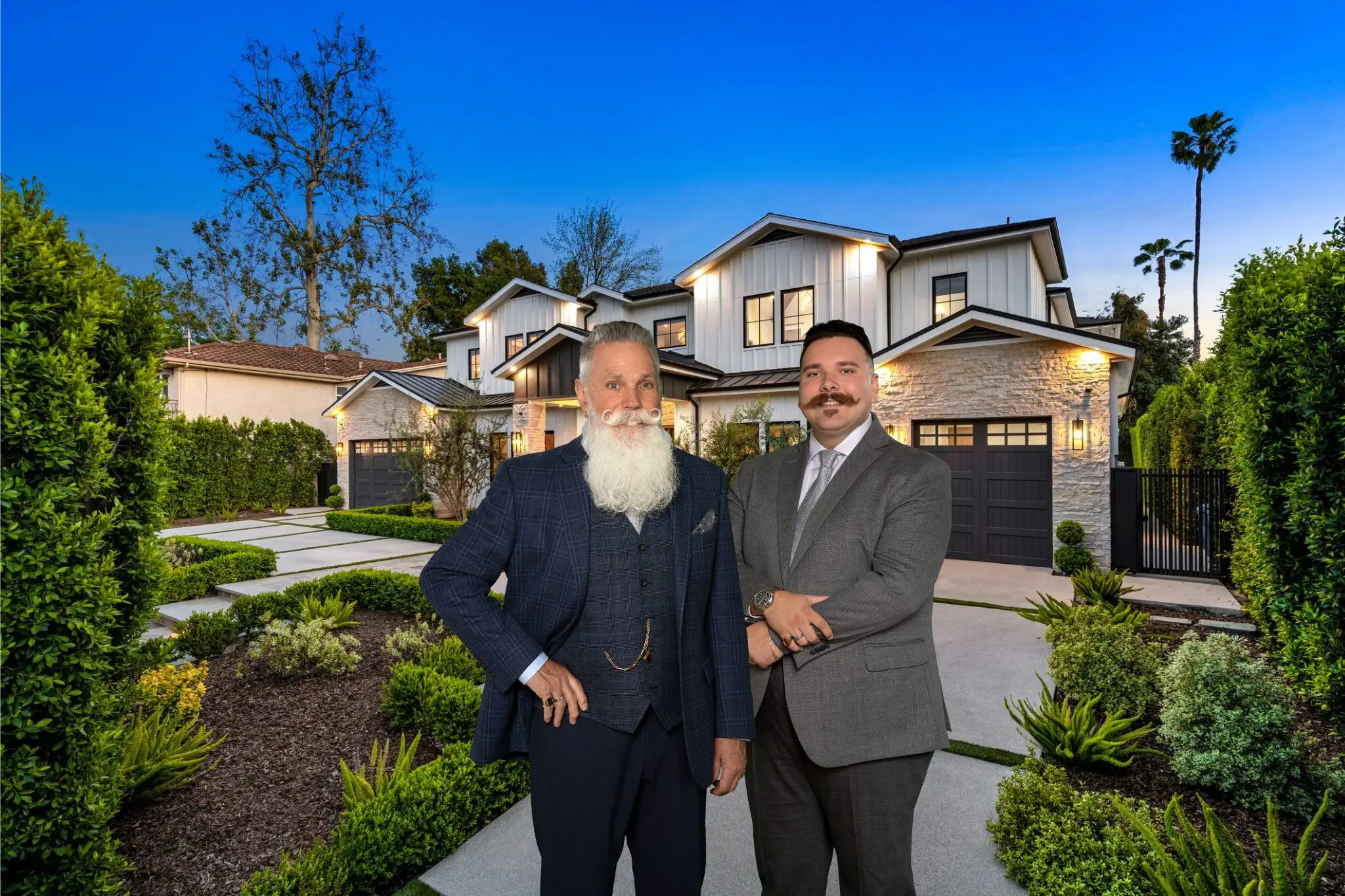 Two men in suits posing in front of a modern two-story house at dusk.