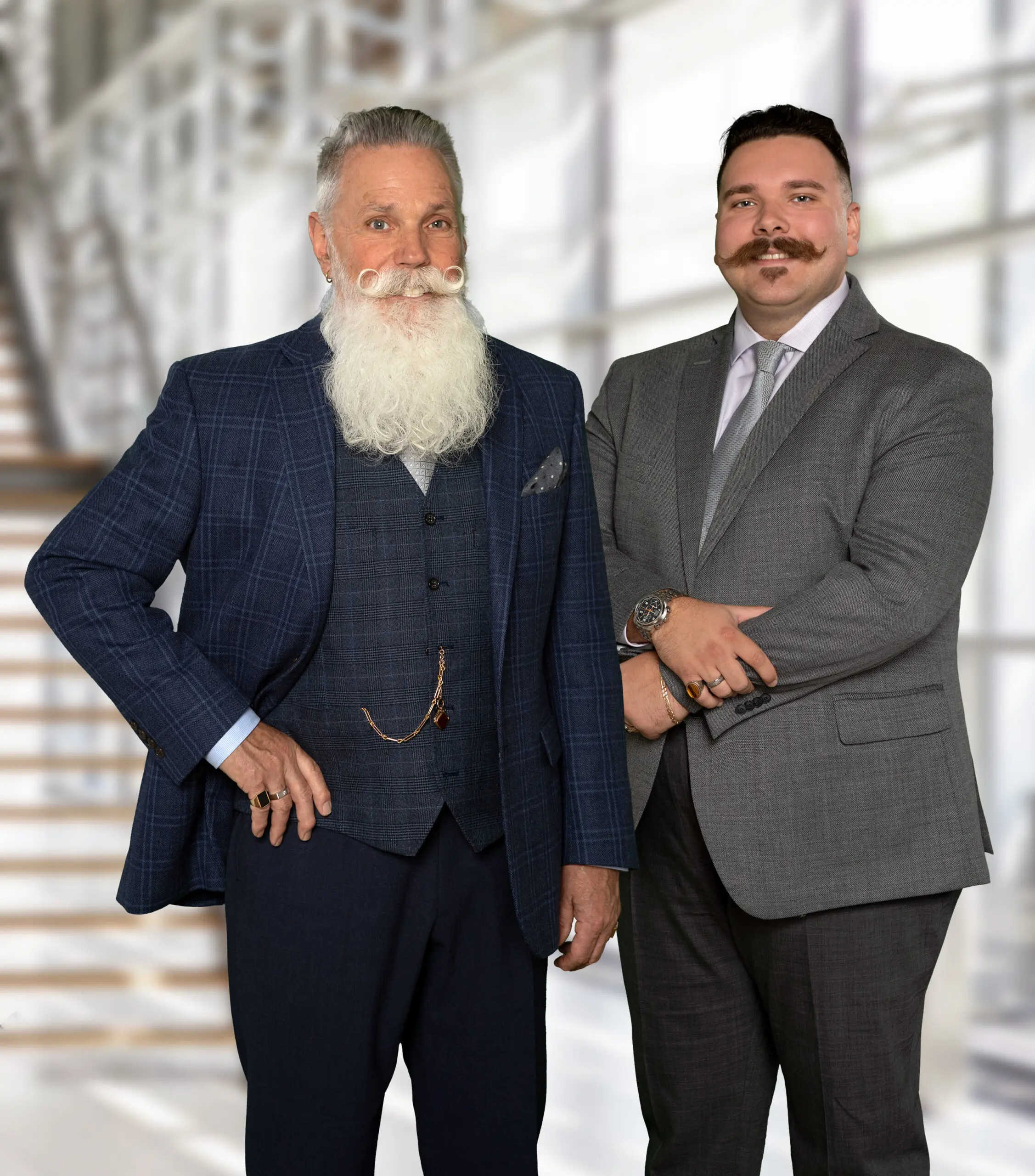 Two men in suits, one with a white beard and the other with a mustache, posing professionally.