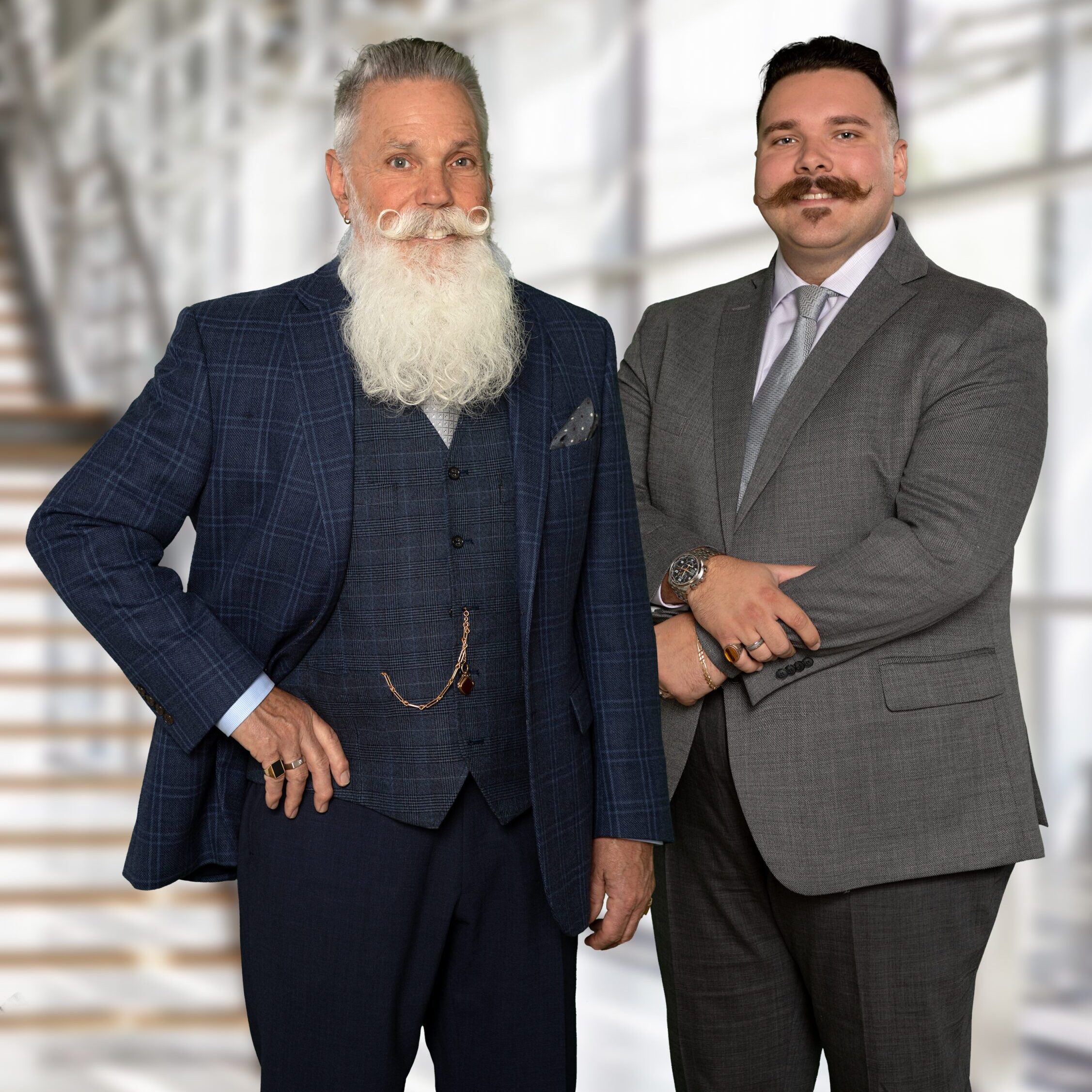 Two men in suits, one with a white beard and the other with a mustache, posing professionally.